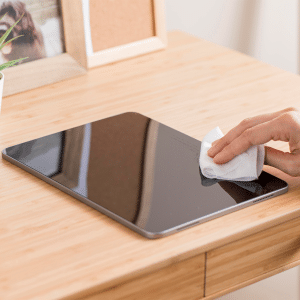 A hand uses a white cloth to give an iPad screen clean on a wooden desk, beside a small potted plant, a framed photo, and a corkboard.