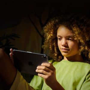 A child with curly hair, wearing a green shirt, sits in a dimly lit room and looks intently at a tablet, checking a Family member purchase made through Apple Family Sharing. Warm light highlights the child’s face and hair.