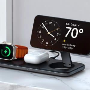 A wireless charging station on a marble surface holds a smartwatch, wireless earbuds, and a smartphone displaying iPhone StandBy with the clock and San Diego weather (70°F, mostly sunny), charging all devices at once.
