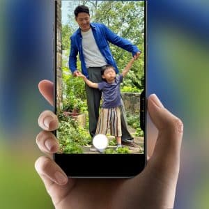 iPhone Video - A hand holds a smartphone displaying an iPhone Video being taken of a smiling man in a blue jacket holding hands with a young child in a striped skirt, standing together outdoors in a lush garden.