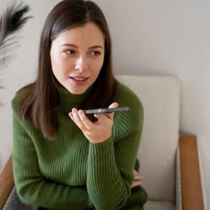A young woman with brown hair in a green sweater sits on a chair, holding an iPhone voice recorder and speaking into it. A peacock feather decorates the background against a plain wall.