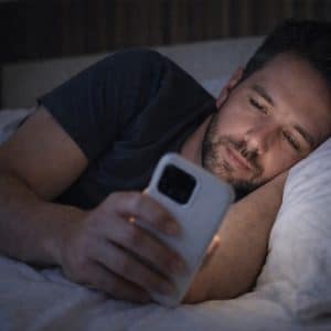 A man lies in bed under a white blanket, propped up on one arm, looking thoughtfully at his iPhone screen in the dim room—perhaps checking Night Shift settings to ease his eyes.