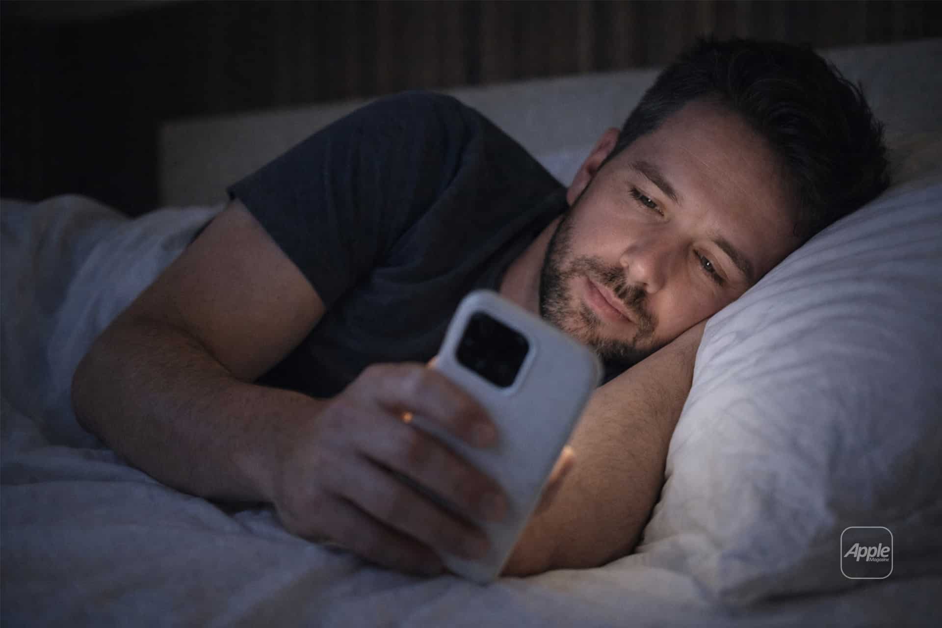 A man lies in bed under a white blanket, propped up on one arm, looking thoughtfully at his iPhone screen in the dim room—perhaps checking Night Shift settings to ease his eyes.