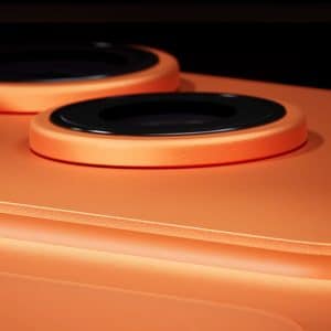 Close-up view of an orange smartphone's rear camera module, featuring three raised camera lenses and a small black sensor set against a smooth matte surface with an iPhone aluminium frame. - iPhone 17 Pro Orange