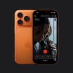 An orange iPhone with an aluminium frame and three rear cameras is shown, its screen displaying a video recording interface capturing a close-up of a person playing a cello against a dark background.