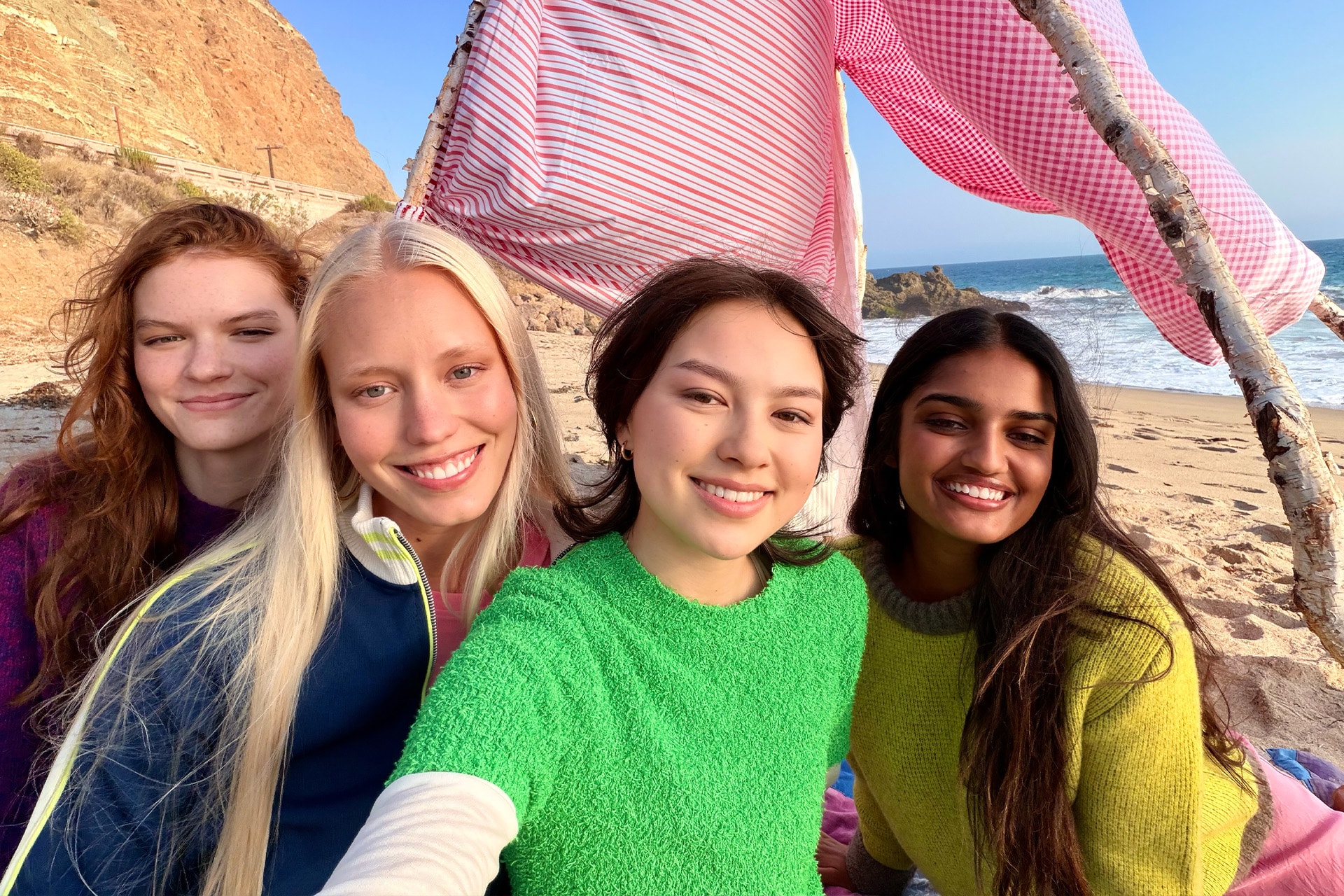 Four young women smile and pose for a selfie on a sandy beach with the ocean and cliffs in the background. They sit under a striped pink-and-white canopy made with sticks, capturing the sunny day in fun iPhone Photos.