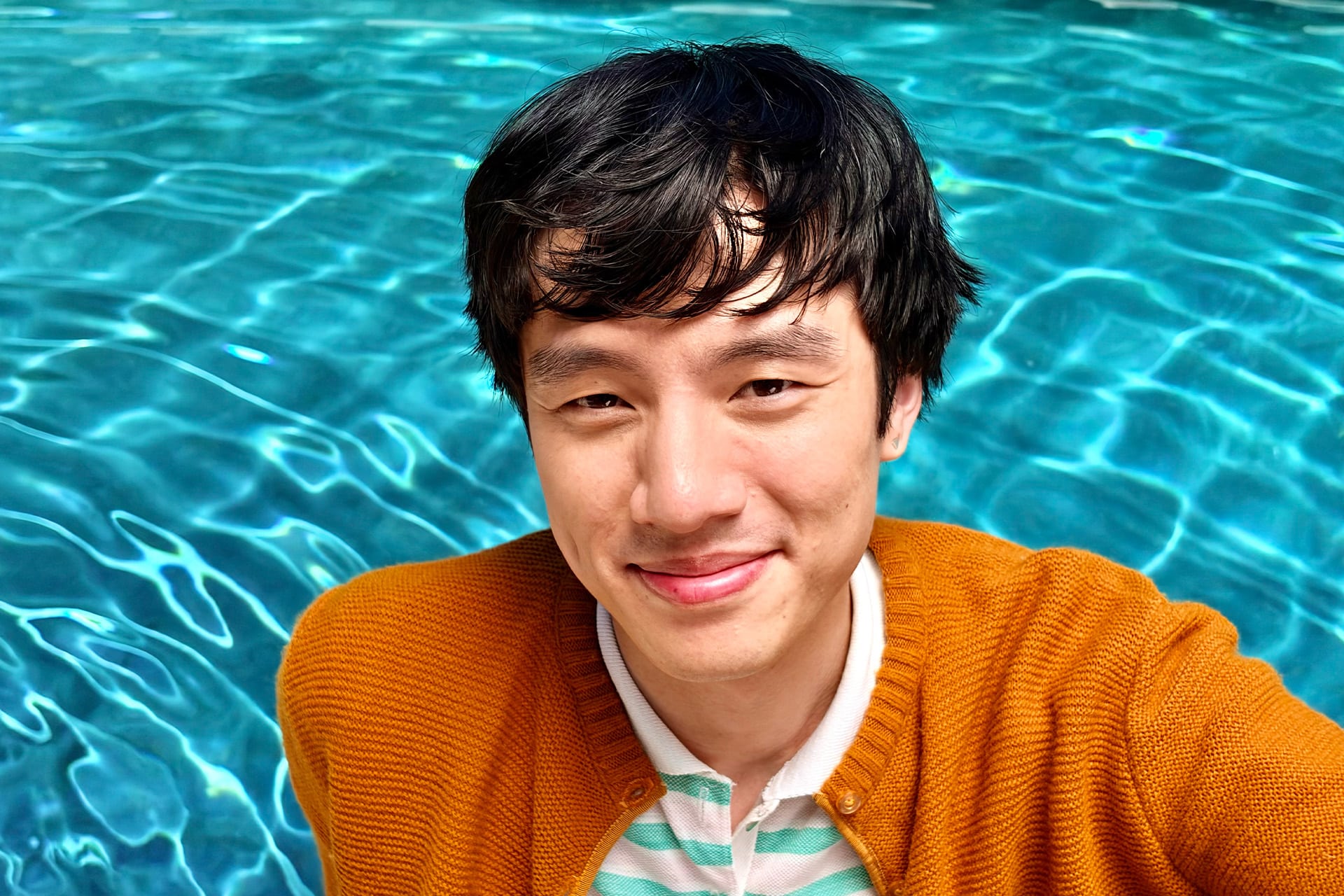 A person with short dark hair smiles at the camera for an iPhone photo, wearing an orange sweater over a striped shirt, standing in front of a swimming pool with clear blue water.