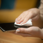 A person is performing an iPhone Clean, gently wiping the smartphone screen with a soft white cloth, holding the phone in one hand on a wooden surface background.