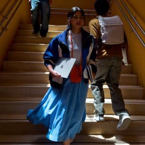 A young woman in a blue skirt and jacket holds her First Mac laptop while walking down sunlit stairs, with two people ahead of her, one carrying a backpack. The setting is indoors, with yellow walls and metal railings.