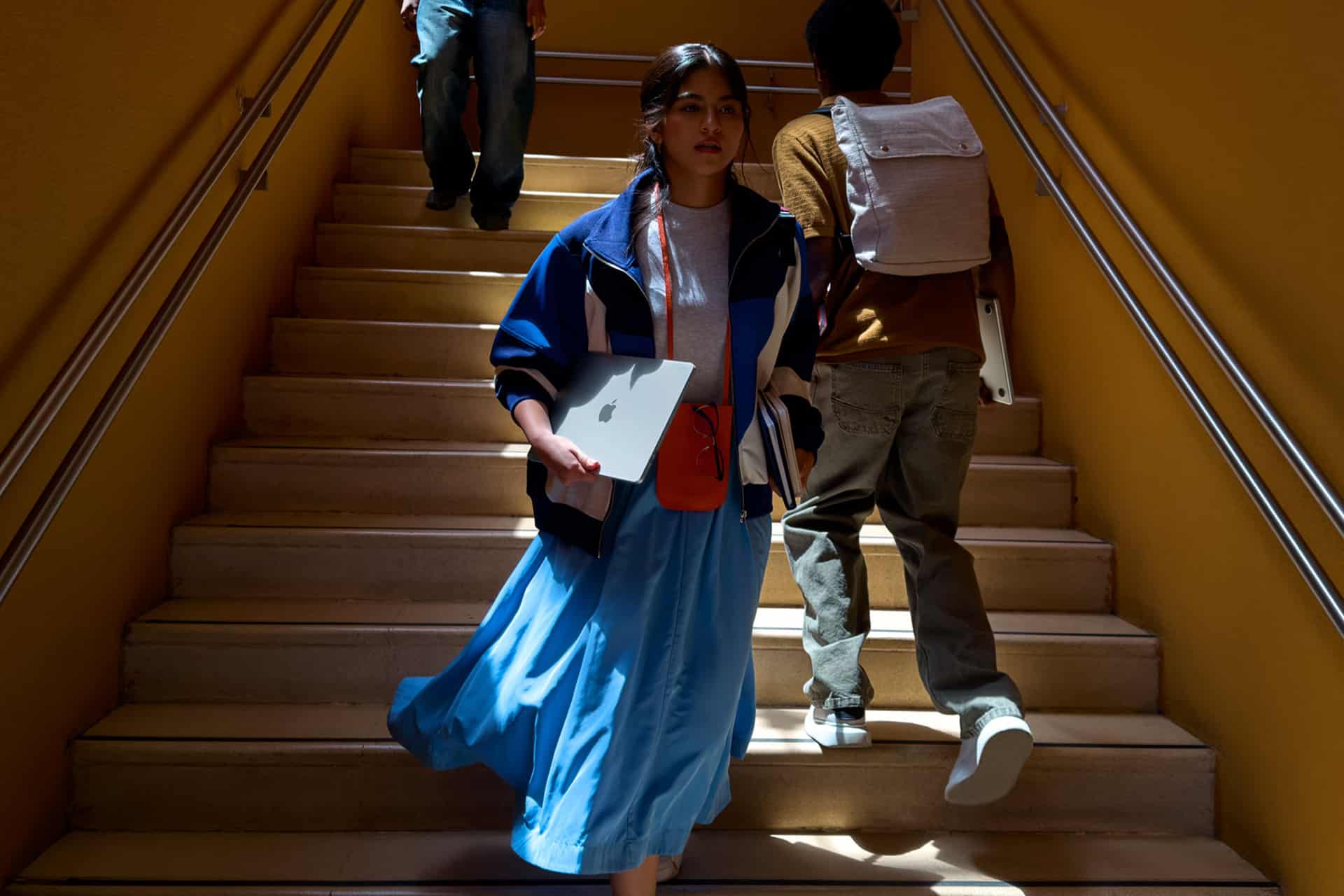 A young woman in a blue skirt and jacket holds her First Mac laptop while walking down sunlit stairs, with two people ahead of her, one carrying a backpack. The setting is indoors, with yellow walls and metal railings.