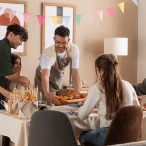 A group of five people gather around a dining table, smiling and enjoying a meal together. Colorful bunting decorates the room, while an Apple Music guest playlist adds to the festive and warm atmosphere.