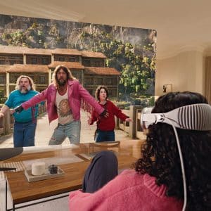 Four people wearing virtual reality headsets sit at a table, experiencing a lifelike, immersive scene powered by visionOS 26 Beta, with three characters running from a large, blocky, video game-style mansion projected in front of them.