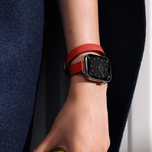 A close-up of a person's hand wearing an Apple Watch with a square black face and a double-wrapped red leather band, highlighting how the Apple Watch Band complements their skin tone, set against a background of dark blue fabric.