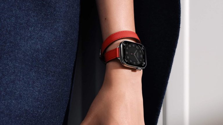 A close-up of a person's hand wearing an Apple Watch with a square black face and a double-wrapped red leather band, highlighting how the Apple Watch Band complements their skin tone, set against a background of dark blue fabric.