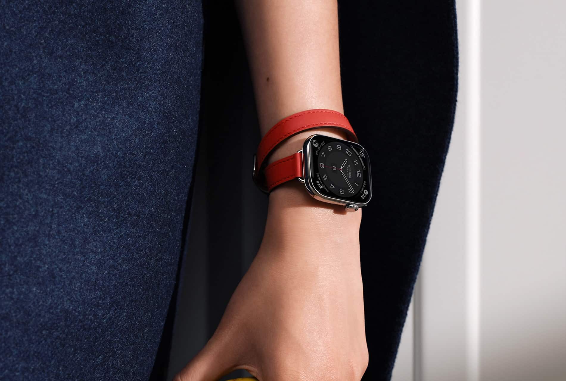 A close-up of a person's hand wearing an Apple Watch with a square black face and a double-wrapped red leather band, highlighting how the Apple Watch Band complements their skin tone, set against a background of dark blue fabric.