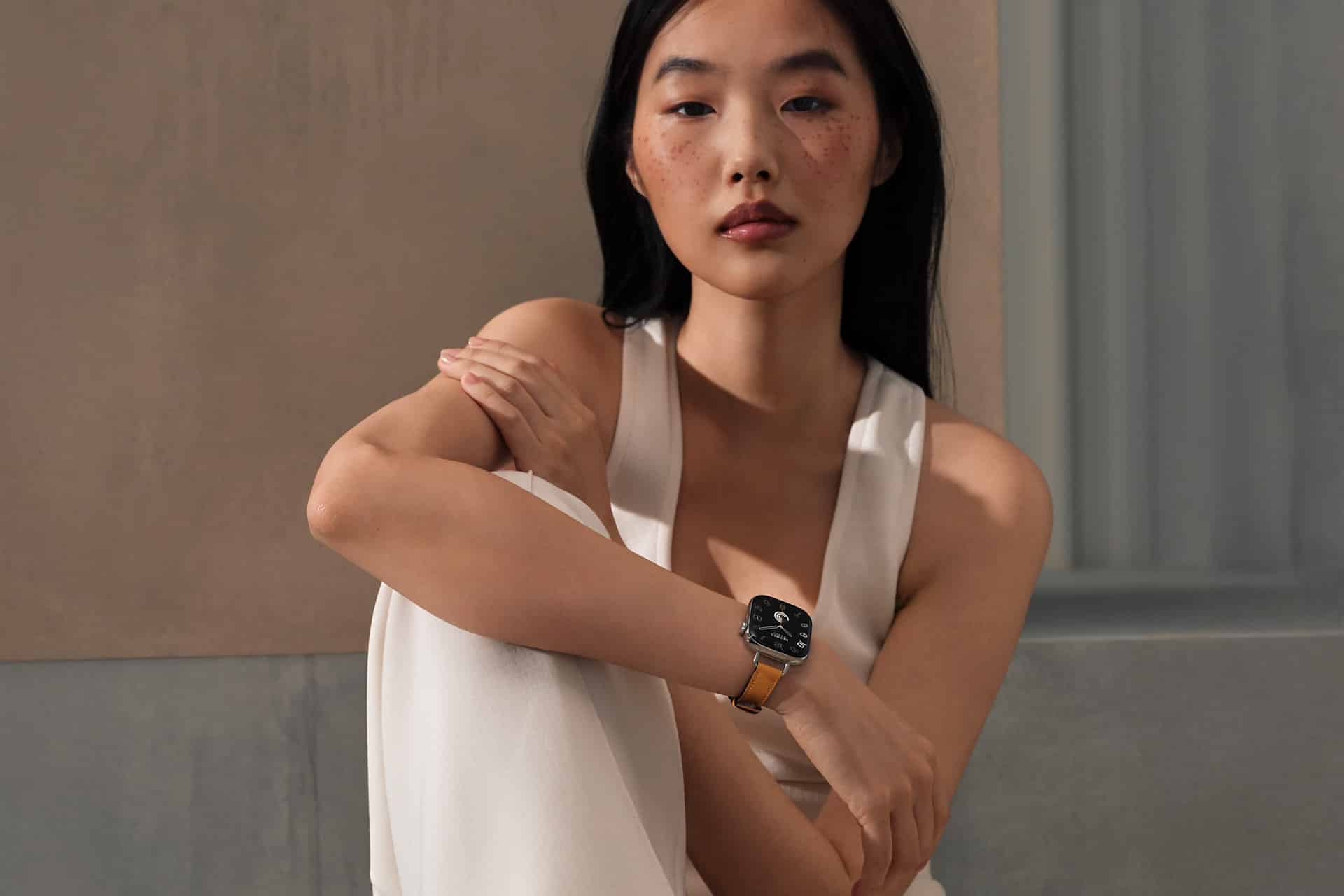 A woman with long black hair sits with her arm resting on her knee, wearing a sleeveless white outfit and an Apple Watch Band Skin Tone that complements her look. She gazes thoughtfully at the camera against a neutral indoor background.