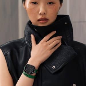 A person with short hair and freckles wears a sleeveless black leather jacket with a high collar and an Apple Watch band in a skin tone shade, posing with one hand touching the collar.