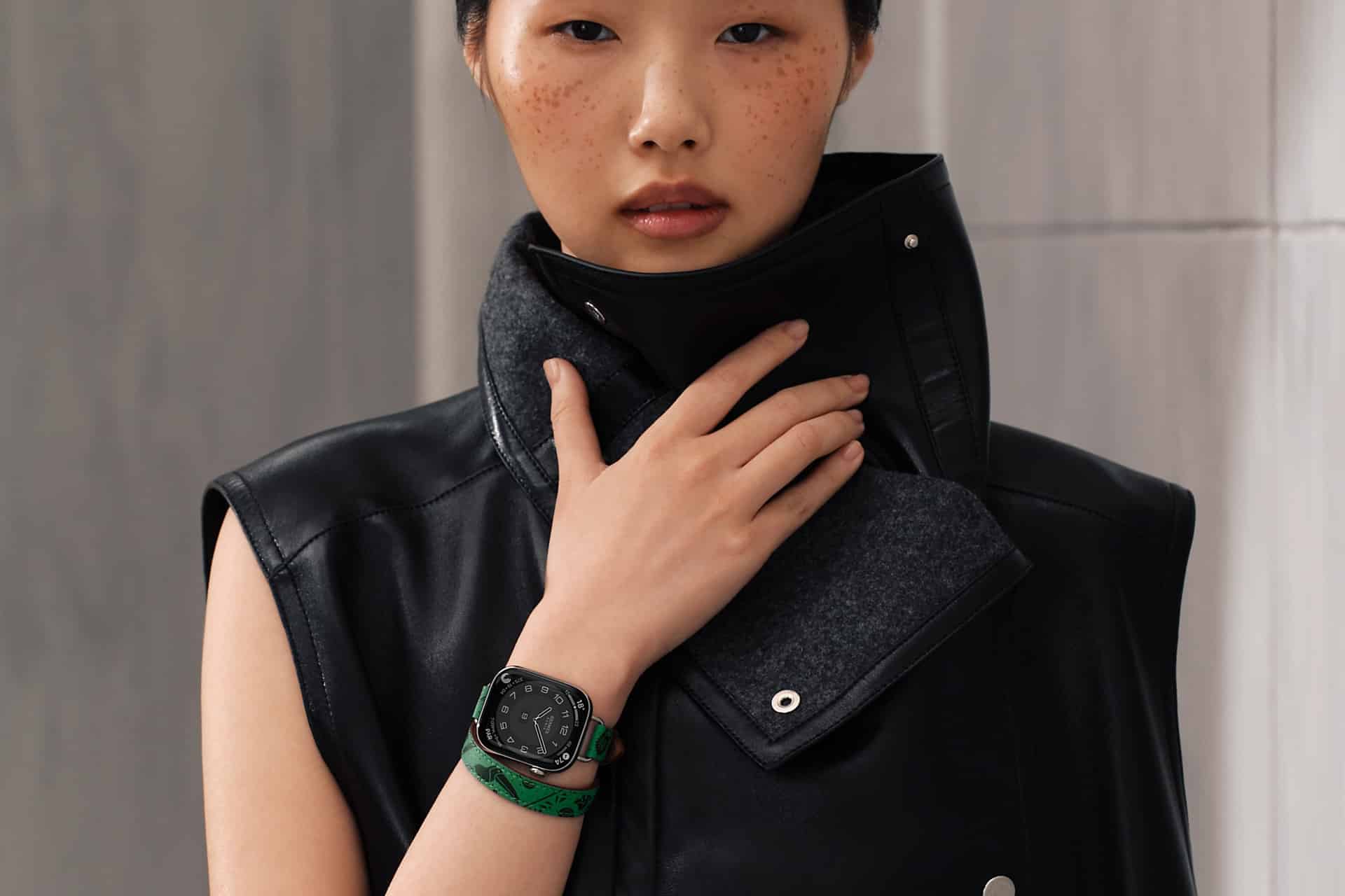 A person with short hair and freckles wears a sleeveless black leather jacket with a high collar and an Apple Watch band in a skin tone shade, posing with one hand touching the collar.