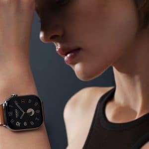 A person wearing a sleeveless black top holds their arm up, showcasing a stylish smartwatch with a square black face and an Apple Watch band in a skin tone shade. The blurred background keeps focus on the watch and the person's profile.