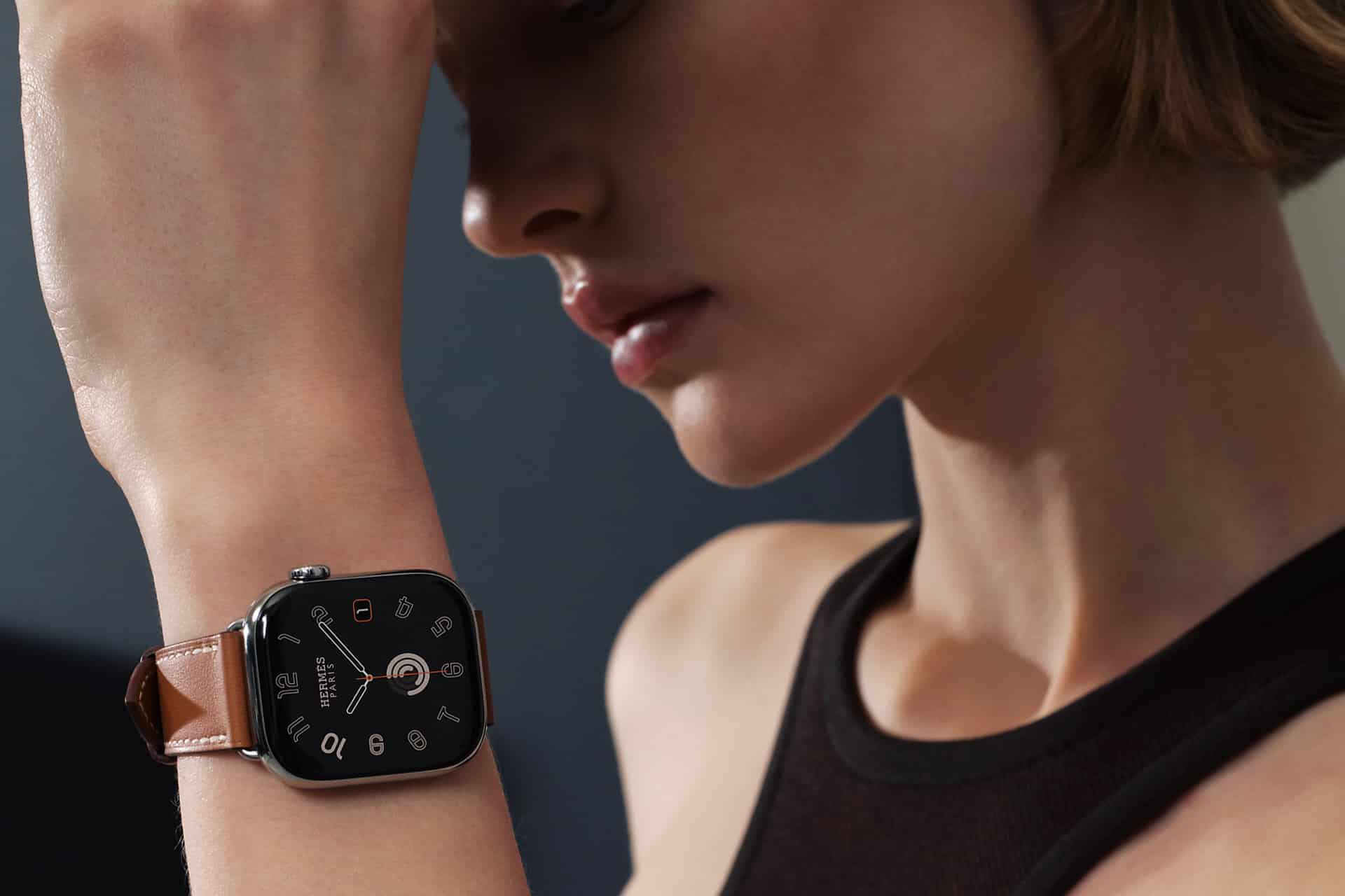 A person wearing a sleeveless black top holds their arm up, showcasing a stylish smartwatch with a square black face and an Apple Watch band in a skin tone shade. The blurred background keeps focus on the watch and the person's profile.