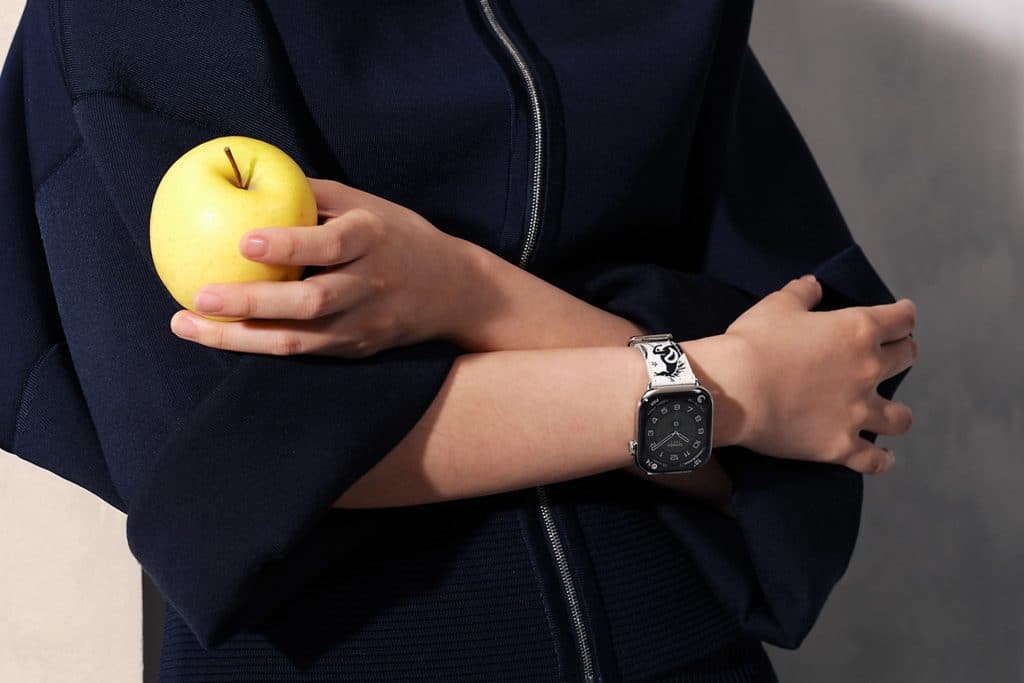 A person in a dark blue outfit holds a yellow apple and folds their arms, showing off a black Apple Watch with a silver band that complements their skin tone.