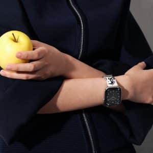 A person in a dark blue outfit holds a yellow apple and folds their arms, showing off a black Apple Watch with a silver band that complements their skin tone.