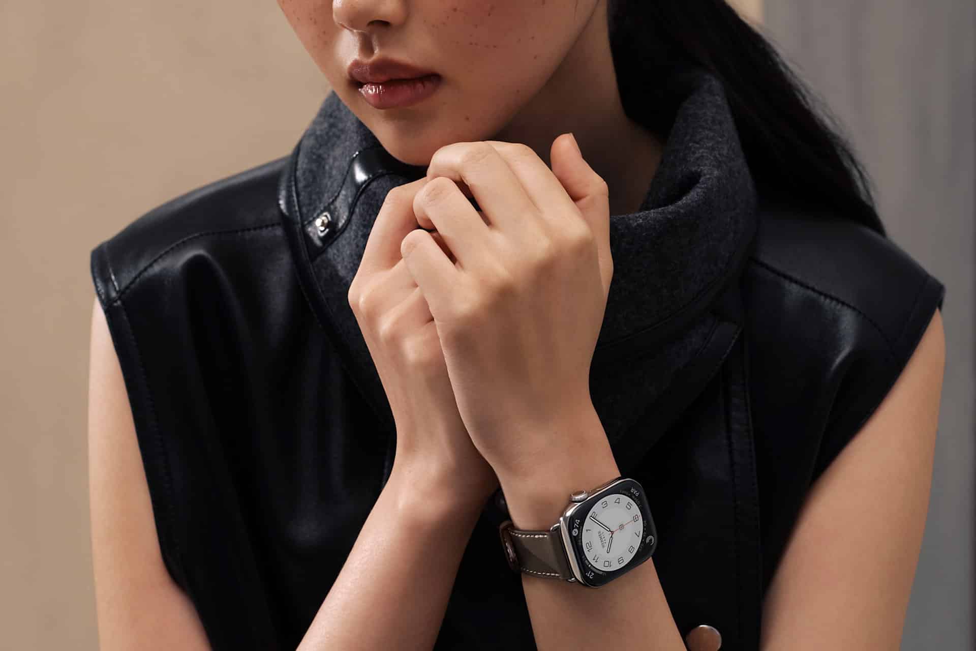A person wearing a sleeveless black leather jacket and a gray scarf holds their hands together near their face, displaying an Apple Watch Band Skin Tone wristwatch with a white round face and a square silver casing.