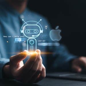 A person holds a glowing holographic chatbot icon with the words “How can I help you?” in front of a laptop, while the Apple logo and references to Apple Siri and AI agents appear in the background on a dark surface.