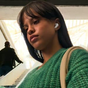 A woman wearing a green sweater and AirPods with Conversation Boost stands on an escalator, holding her phone. People are visible in the background, and natural light pours through a large glass ceiling.