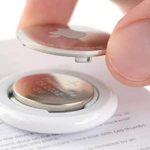 A close-up of a hand opening the back cover of an Apple device, revealing an AirTag battery (CR2032 coin cell) inside the white circular casing. The device rests on top of a printed instruction manual.