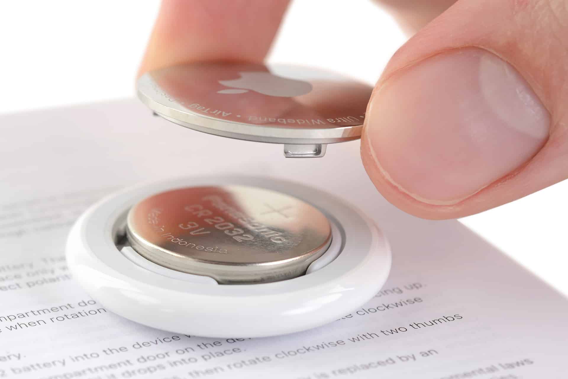 A close-up of a hand opening the back cover of an Apple device, revealing an AirTag battery (CR2032 coin cell) inside the white circular casing. The device rests on top of a printed instruction manual.