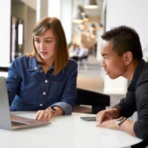 Two people sit at a white table in the bright Apple Cork Office, closely looking at a laptop screen. One gestures toward the laptop while the other listens attentively, reflecting a collaborative atmosphere in this modern workspace.