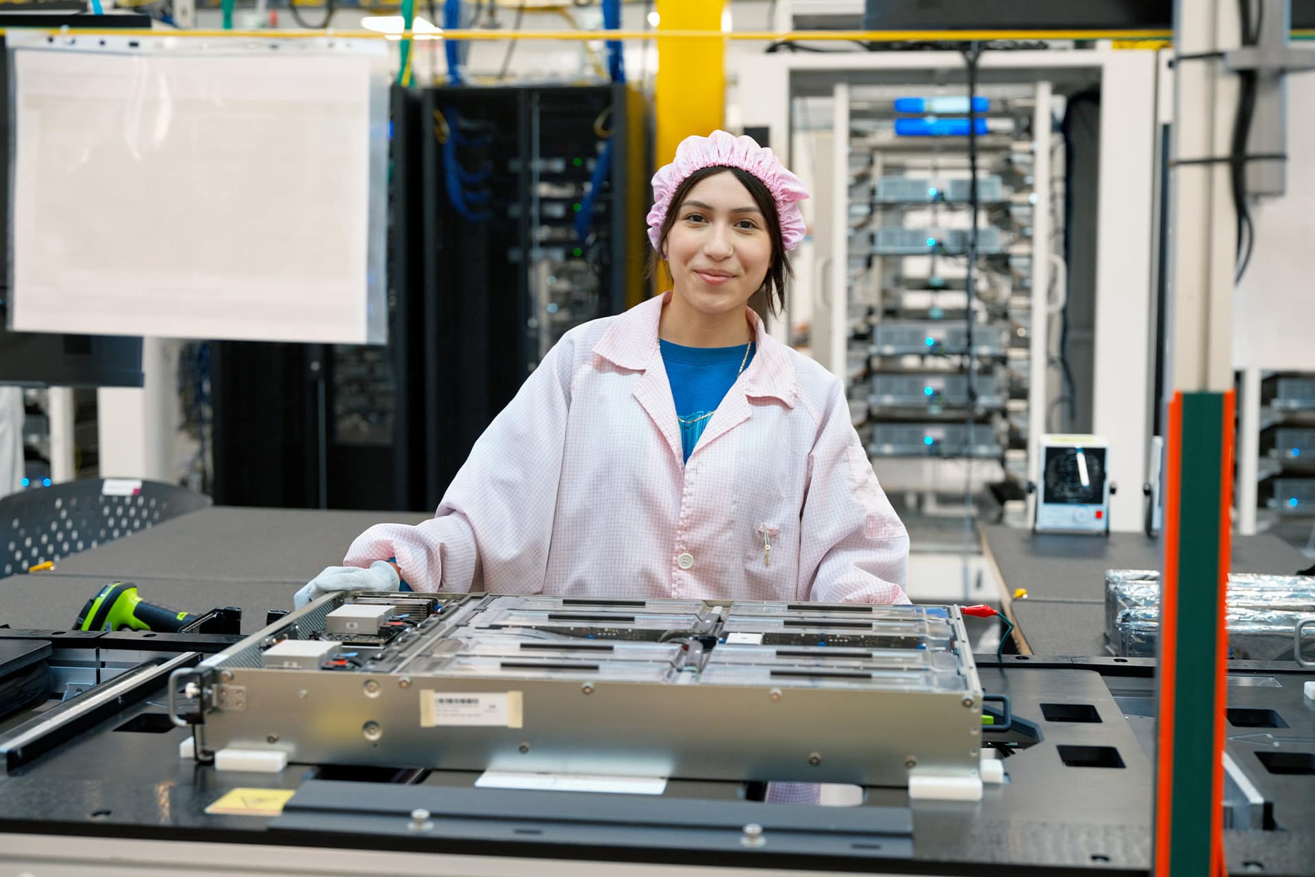 A woman in a pink lab coat and cap works in a high-tech Apple US Production facility, standing behind electronic equipment on an assembly table, with servers and advanced machinery visible in the background.