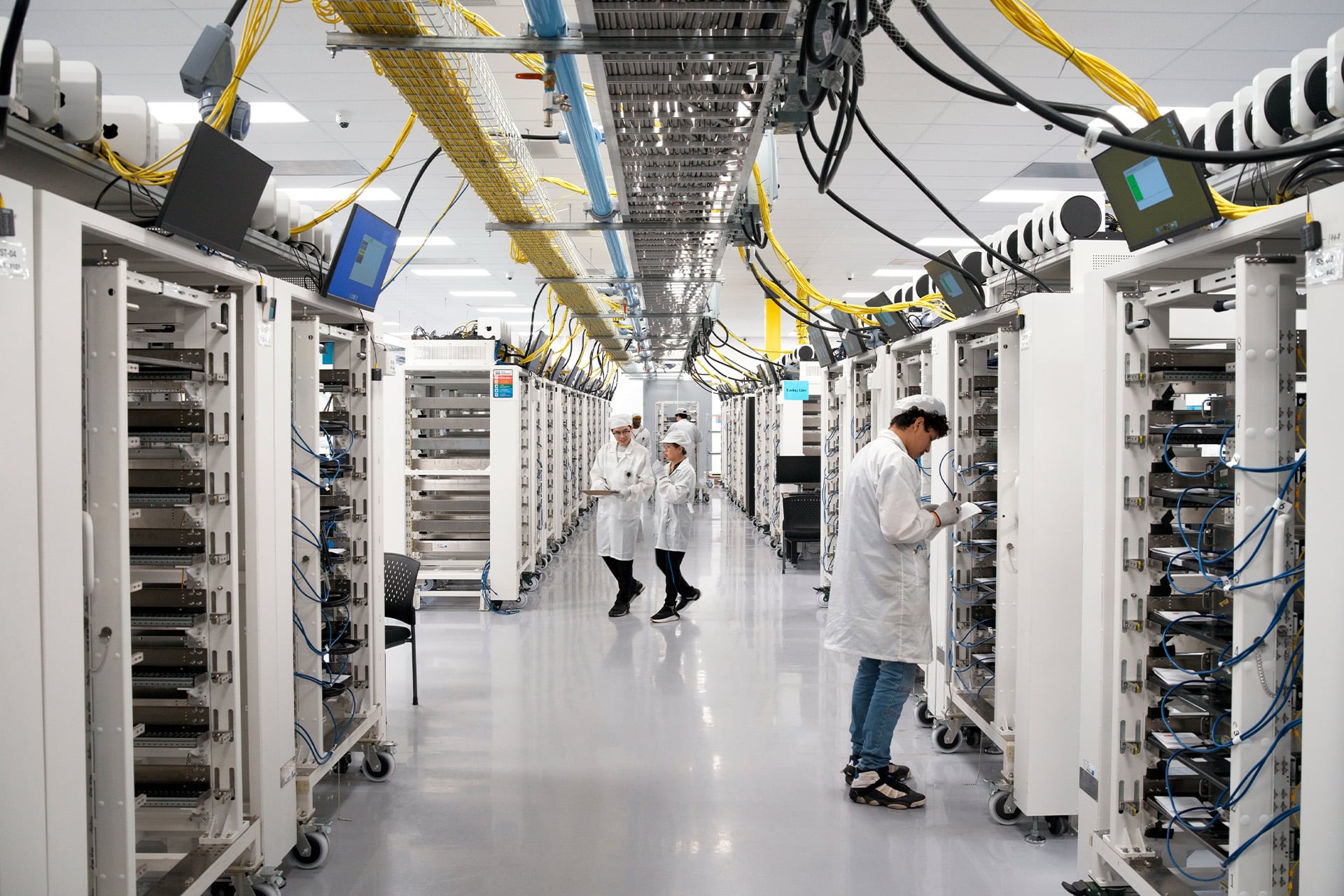 Technicians in white lab coats work among rows of large server racks and cables in a brightly lit data center, monitoring systems and checking equipment as part of Apple US Production operations.