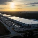 Aerial view of a large industrial warehouse complex, possibly linked to Apple US Production, with long white-roofed buildings, many parked cars at sunset, surrounding roads, trees, and a distant city skyline.