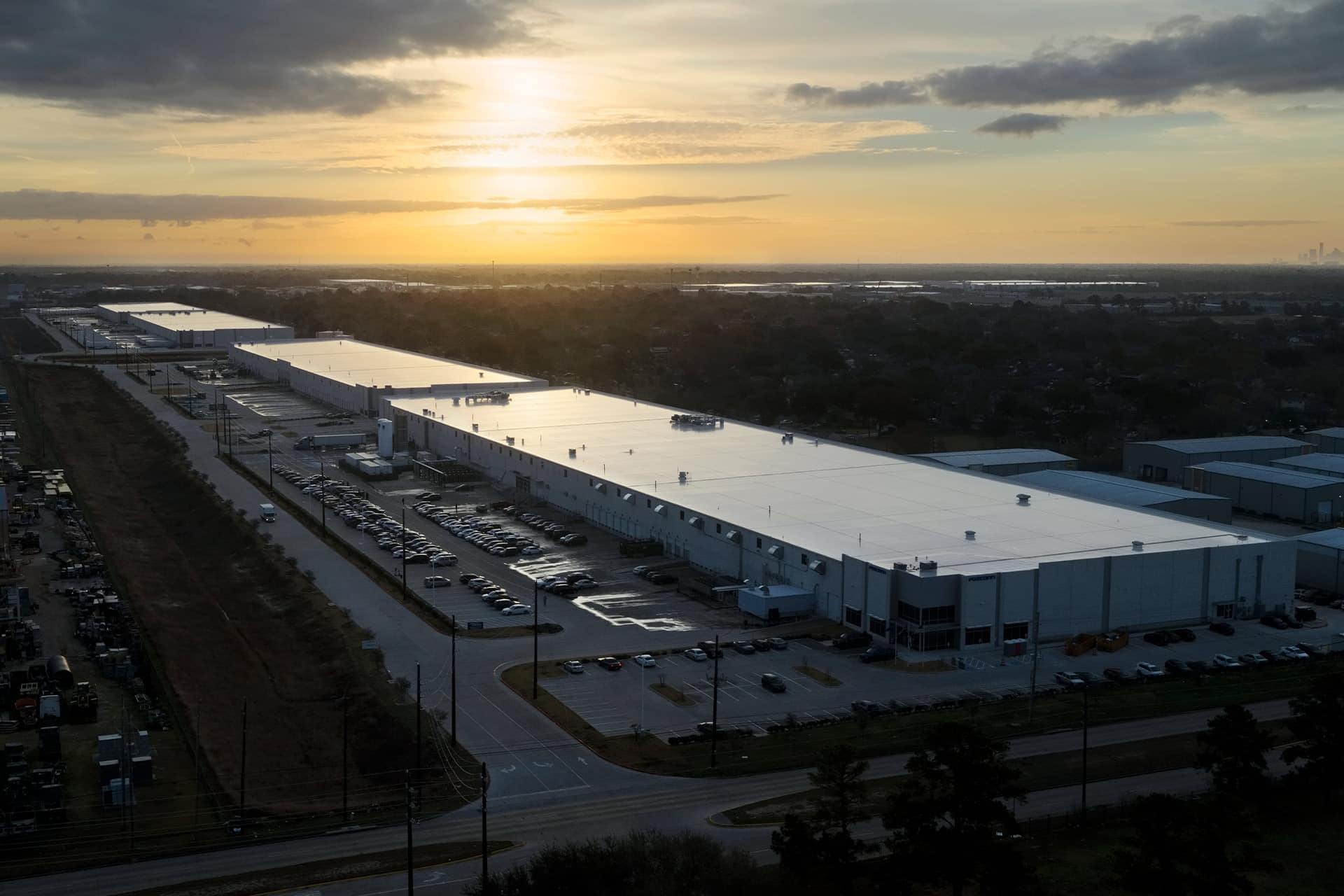 Aerial view of a large industrial warehouse complex, possibly linked to Apple US Production, with long white-roofed buildings, many parked cars at sunset, surrounding roads, trees, and a distant city skyline.
