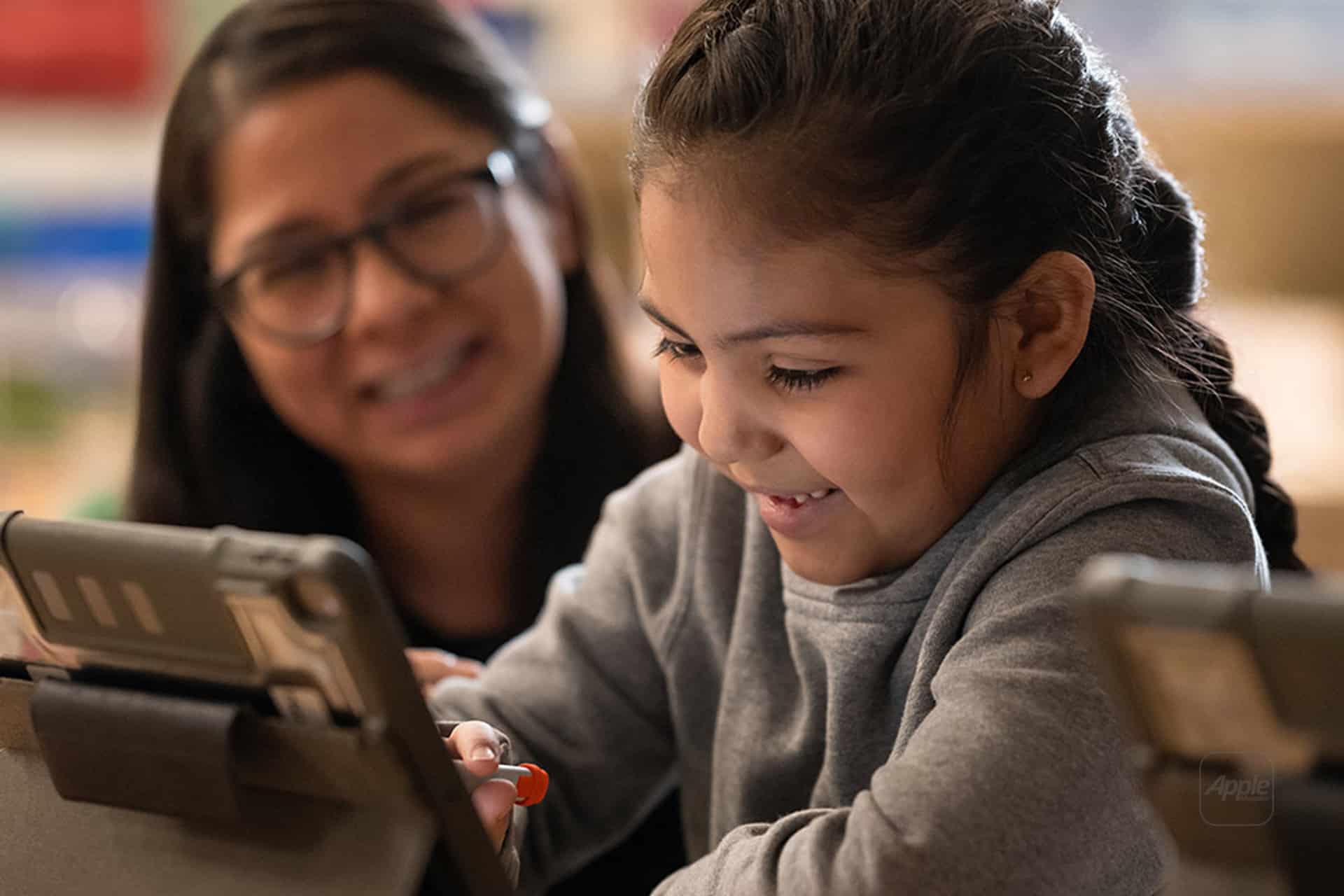 A young girl smiles while using a tablet, with an adult woman sitting beside her, also smiling. Both appear engaged and happy in an Apple Classroom learning environment.