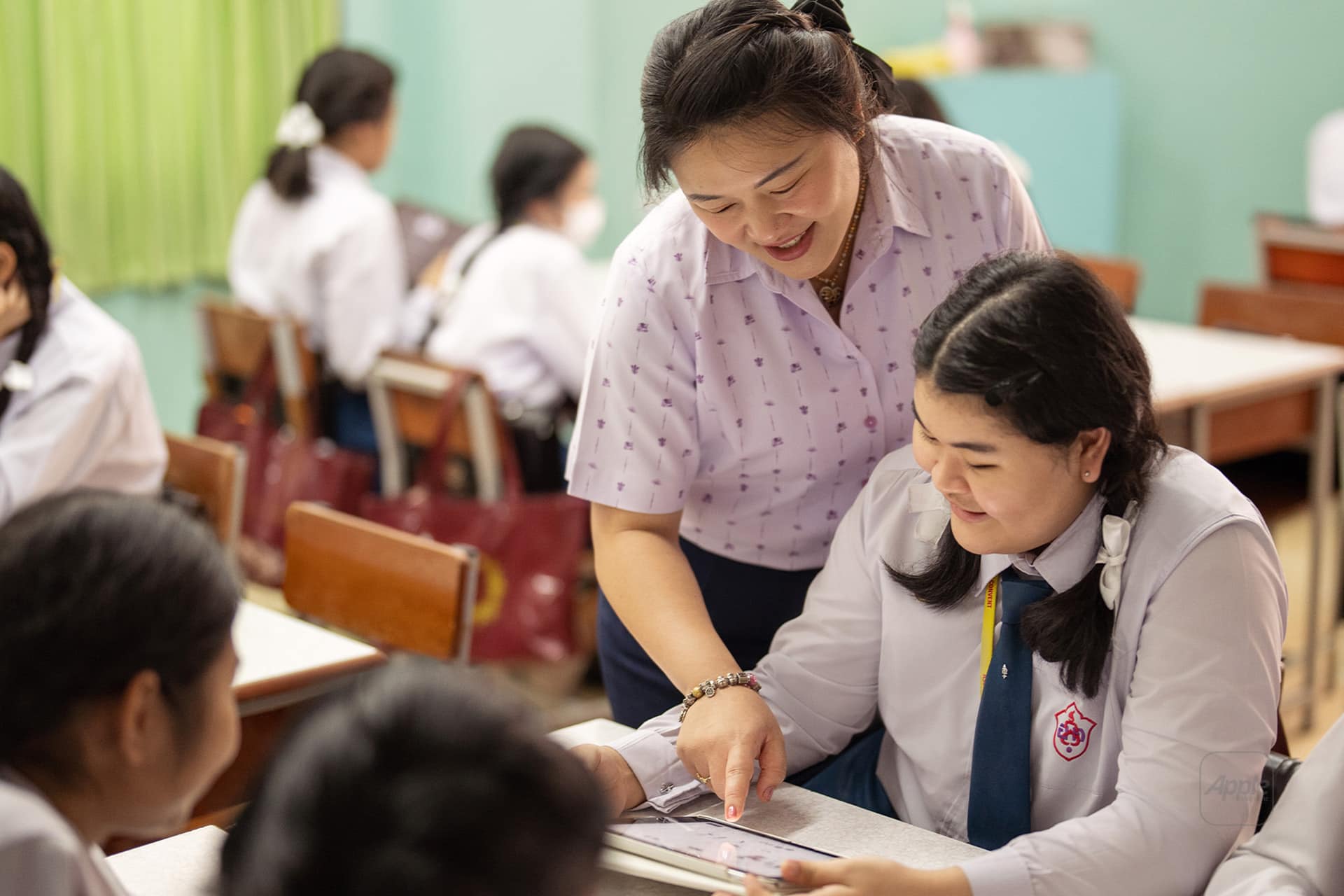 A teacher smiles as she helps a student with her work in an apple classroom. Other students in uniform are seated at desks in the background, focused on their tasks.