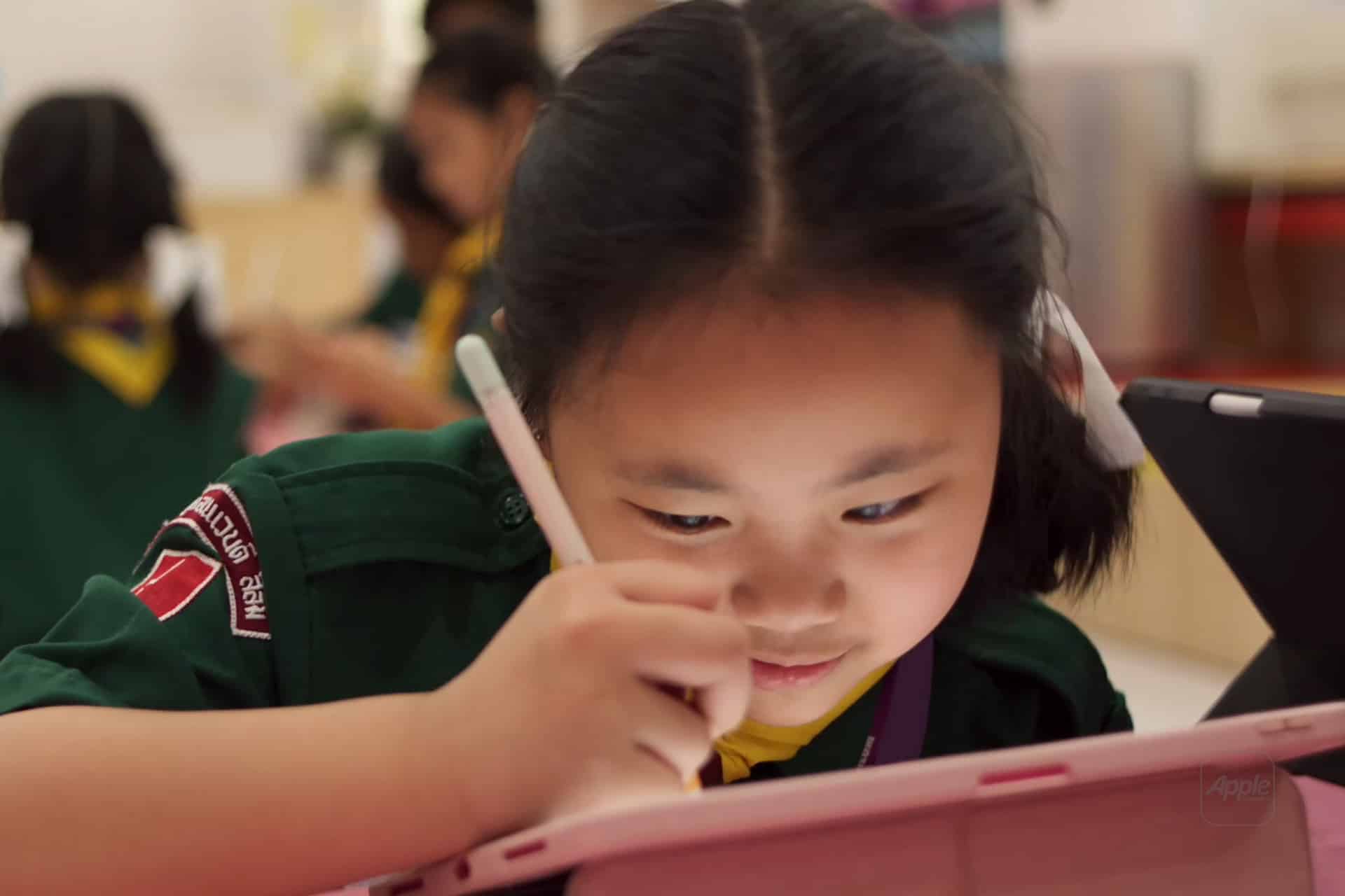 A young girl in a green uniform uses a stylus to draw or write on a pink tablet, focusing intently—immersed in an apple classroom setting. Other children in similar uniforms are blurred in the background.