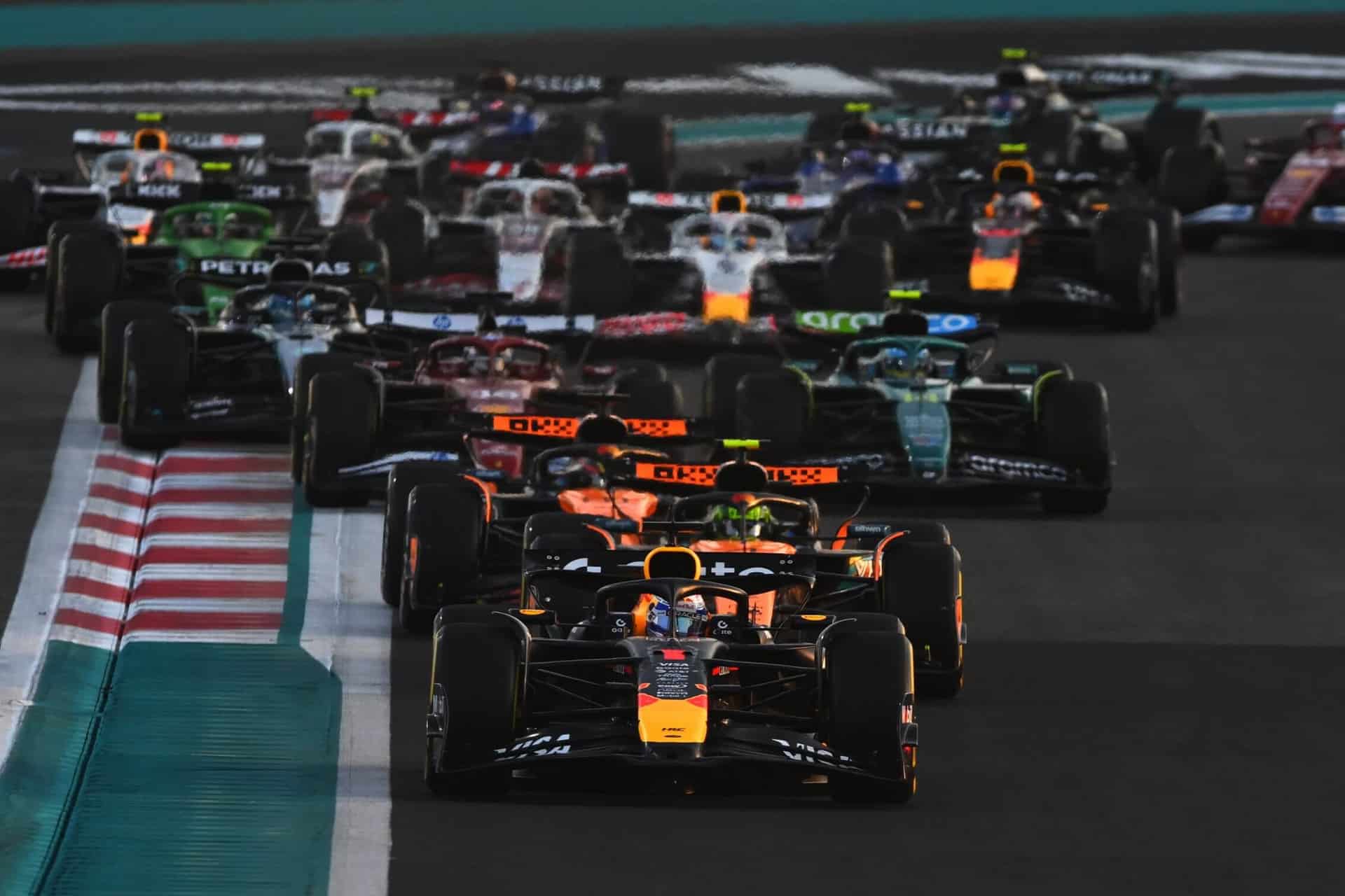 A group of Formula 1 cars race closely together on a track, with the lead cars displaying Red Bull Racing colors. The action-packed scene evokes moments from Netflix's Drive to Survive as the cars turn on red and white curbs.