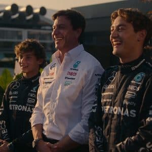 Drive to Survive Season 8 - Three men, two in black Mercedes-AMG Petronas racing suits and one in a white team shirt, stand together smiling outdoors, with greenery and modern buildings in the background—a scene reminiscent of Formula One and Netflix's Drive to Survive.