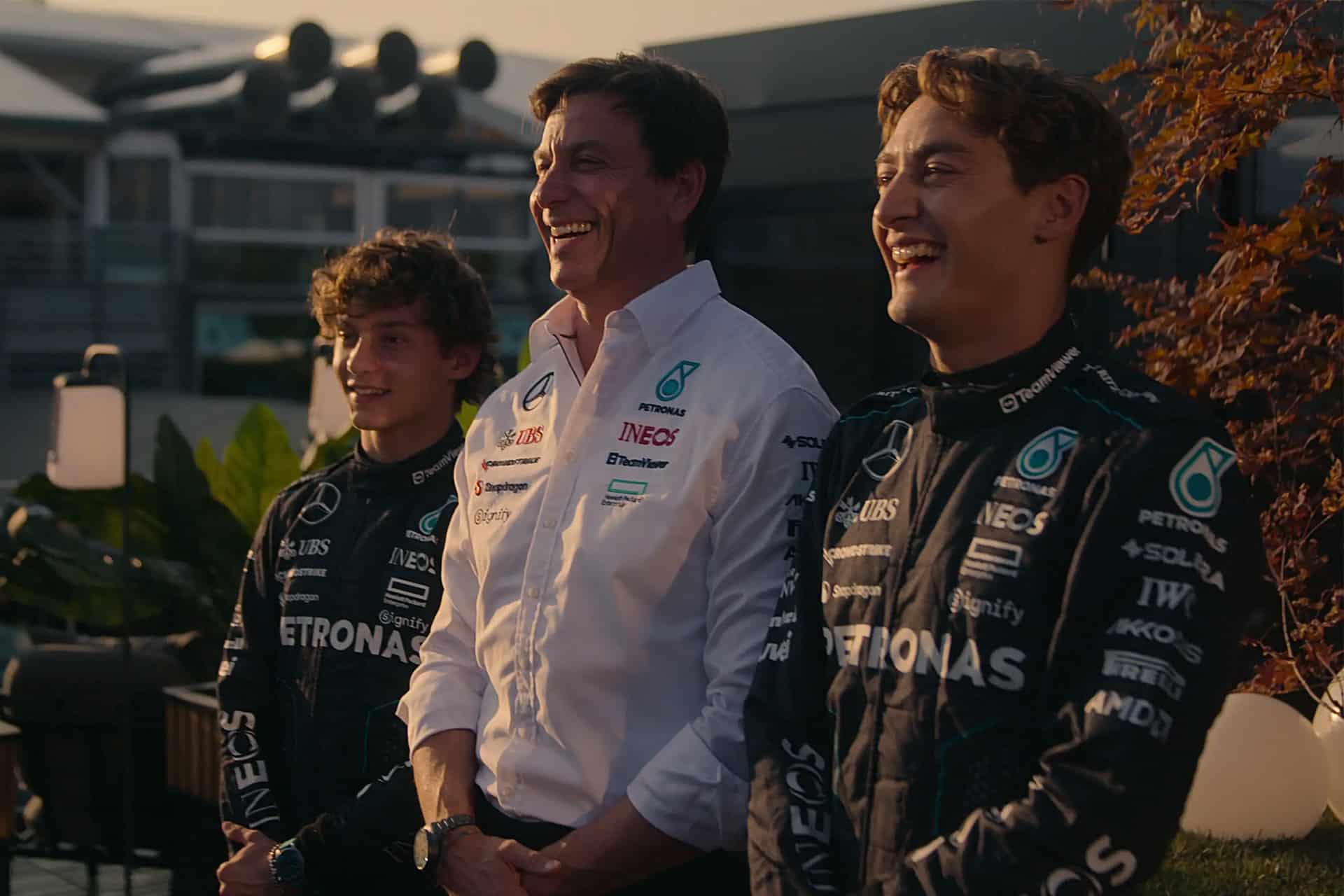 Drive to Survive Season 8 - Three men, two in black Mercedes-AMG Petronas racing suits and one in a white team shirt, stand together smiling outdoors, with greenery and modern buildings in the background—a scene reminiscent of Formula One and Netflix's Drive to Survive.