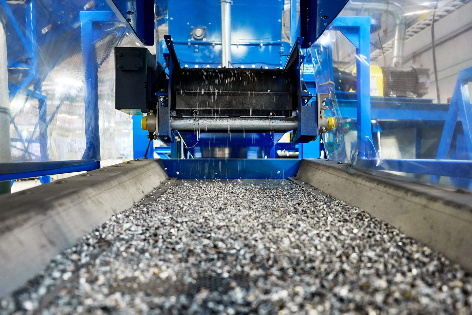 Close-up of a conveyor belt transporting small metal shavings inside an industrial facility, part of the Apple Device Recycling process, with blue machinery and protective barriers visible in the background.