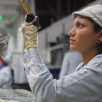 A woman wearing a light-colored uniform, cap, and gloves inspects an object in a factory setting, focusing intently under bright industrial lighting—a scene reflecting Apple sustainability practices in quality control.