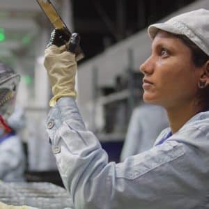 A woman wearing a light-colored uniform, cap, and gloves inspects an object in a factory setting, focusing intently under bright industrial lighting—a scene reflecting Apple sustainability practices in quality control.