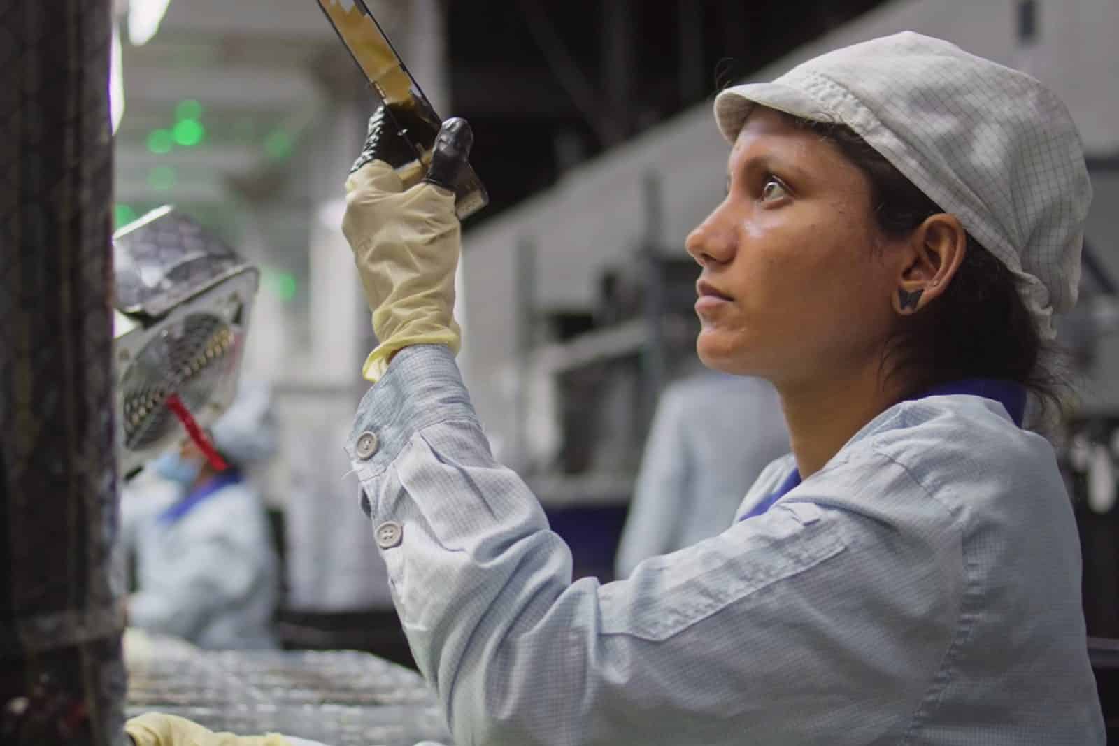 A woman wearing a light-colored uniform, cap, and gloves inspects an object in a factory setting, focusing intently under bright industrial lighting—a scene reflecting Apple sustainability practices in quality control.