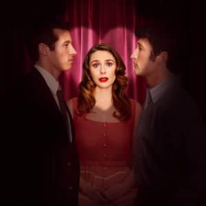 A woman in a red dress stands between two men facing her, with dramatic lighting and a dark red curtain backdrop. She looks forward with a concerned expression while the men gaze at each other in this tense Eternity Movie on Apple TV.
