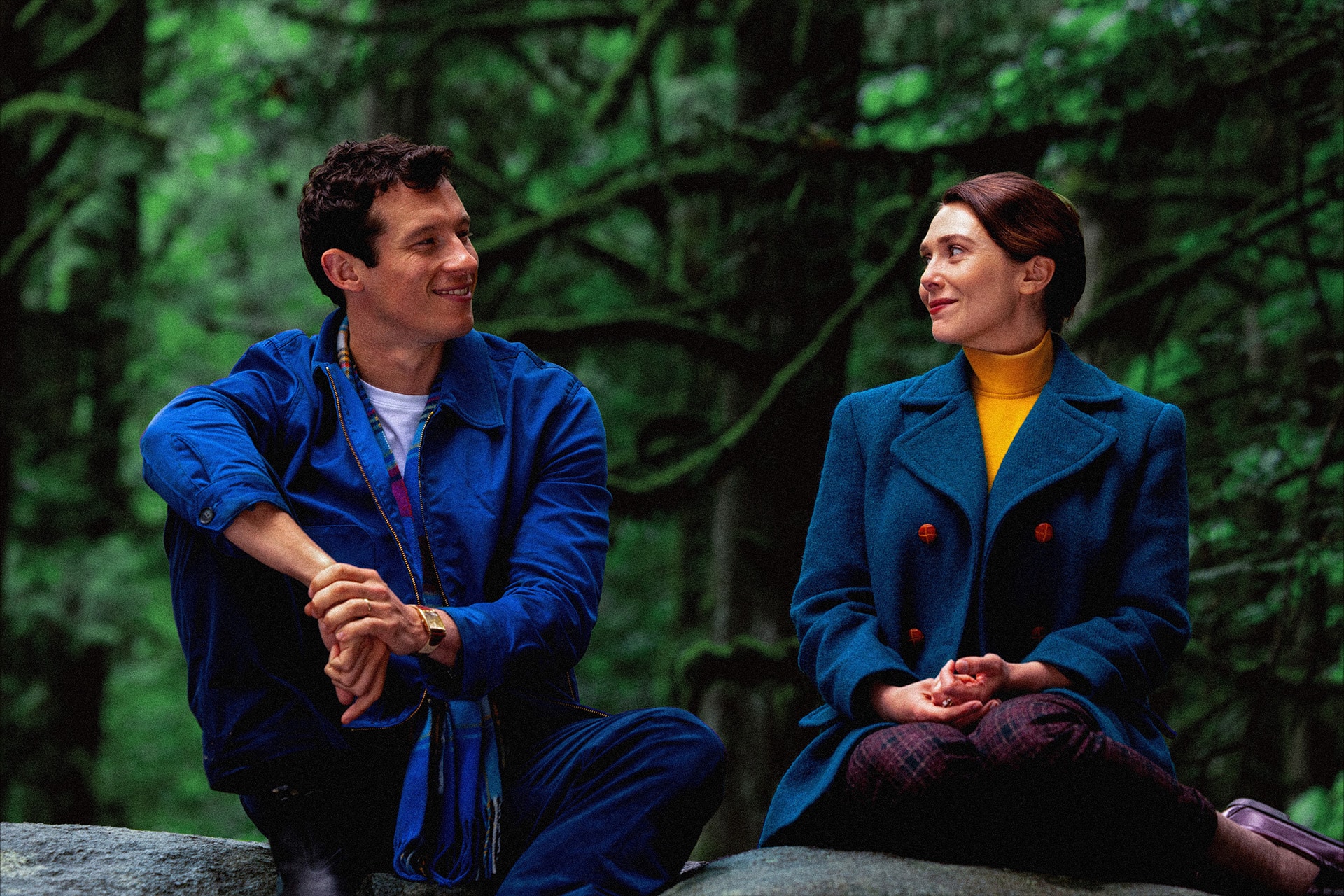 A man and a woman sit on a rock in a lush, green forest, facing each other and smiling warmly—just like a scene from an Apple TV Original. The man wears a blue outfit while the woman dons a teal coat over a yellow turtleneck.