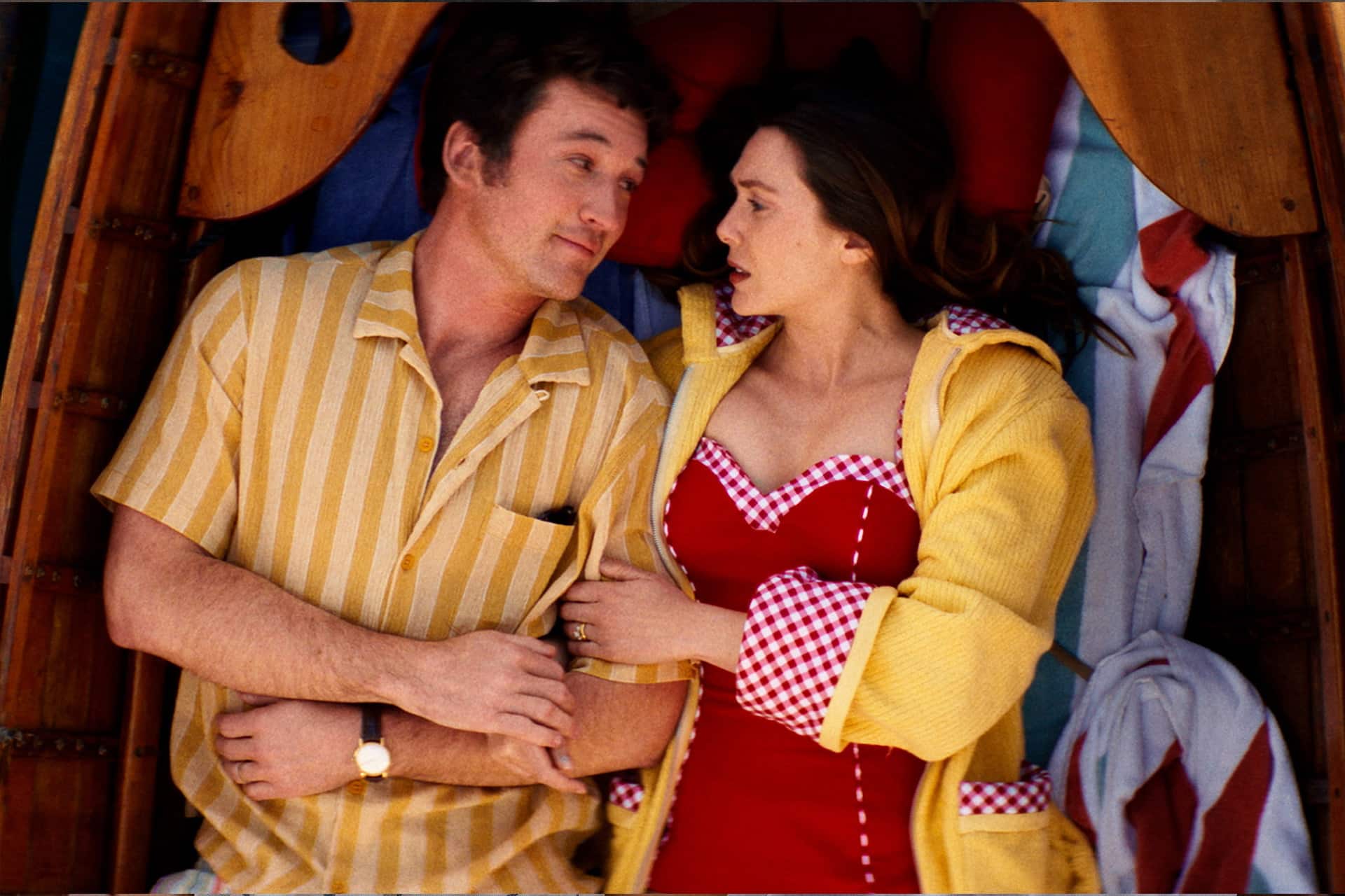 A man in a yellow striped shirt and a woman in a red dress with checkered trim and a yellow robe lie closely together in a wooden boat, looking at each other with affection, as seen in the romantic dramas on Apple TV Originals.