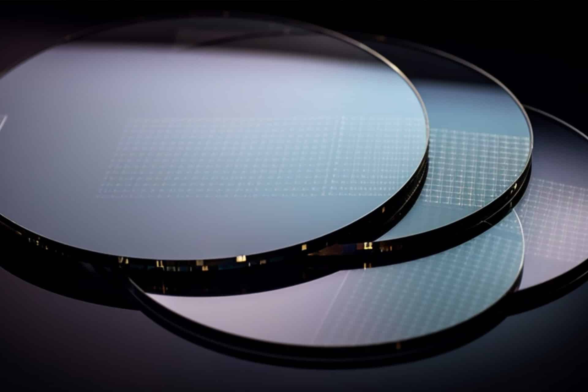 Three thin, reflective silicon wafers with grid patterns—evoking the precision of Apple US Production—rest on a dark, glossy surface, partially overlapped and illuminated by soft, diffused light.